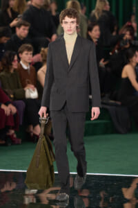 Male model walks a runway in a charcoal gray suit with a beige turtleneck, carrying a large olive-green bag at his side.