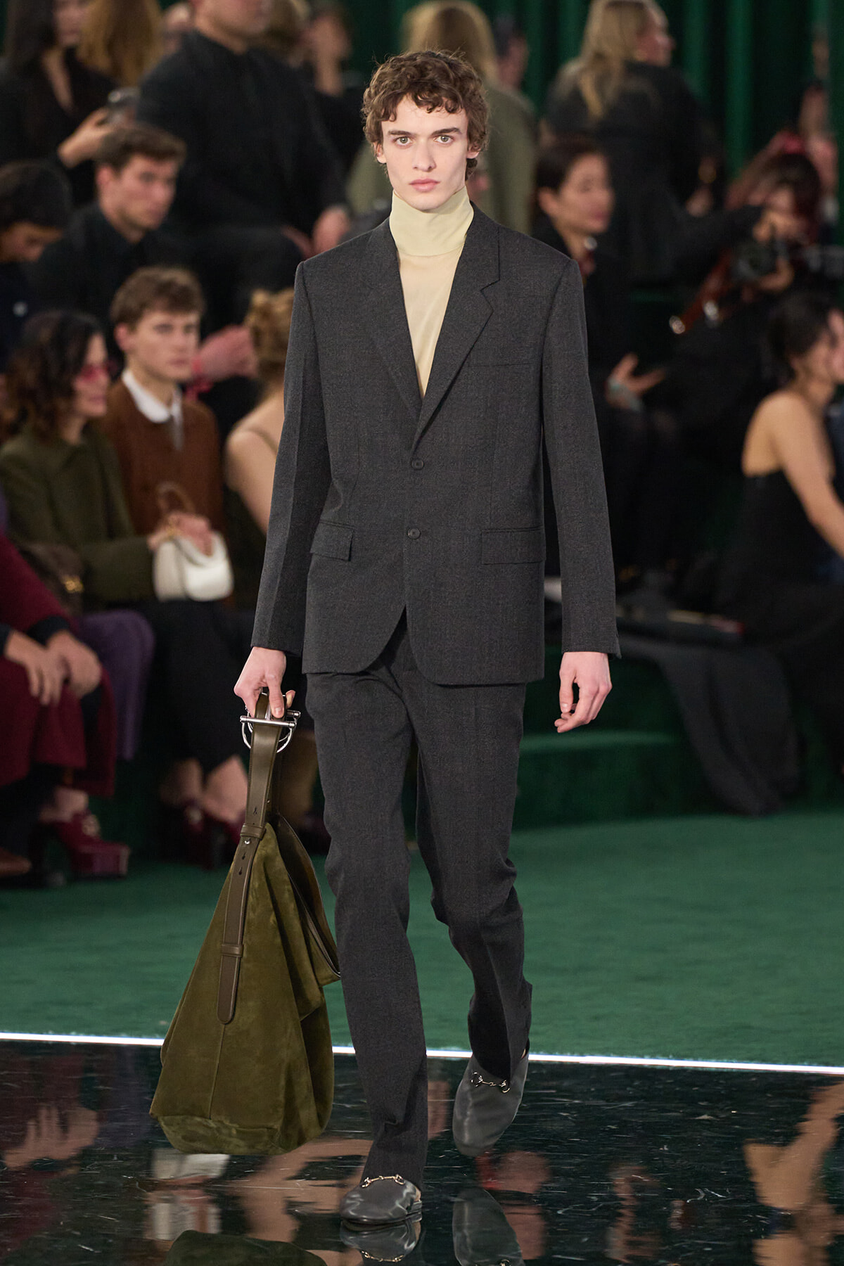 Male model walks a runway in a charcoal gray suit with a beige turtleneck, carrying a large olive-green bag at his side.