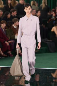 Male model in a pale lavender suit walking the runway, carrying a large beige bag, with spectators in the background.
