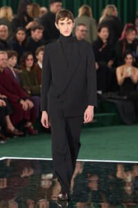 Male model in a black double-breasted suit and turtleneck walks the runway; green carpet and audience behind