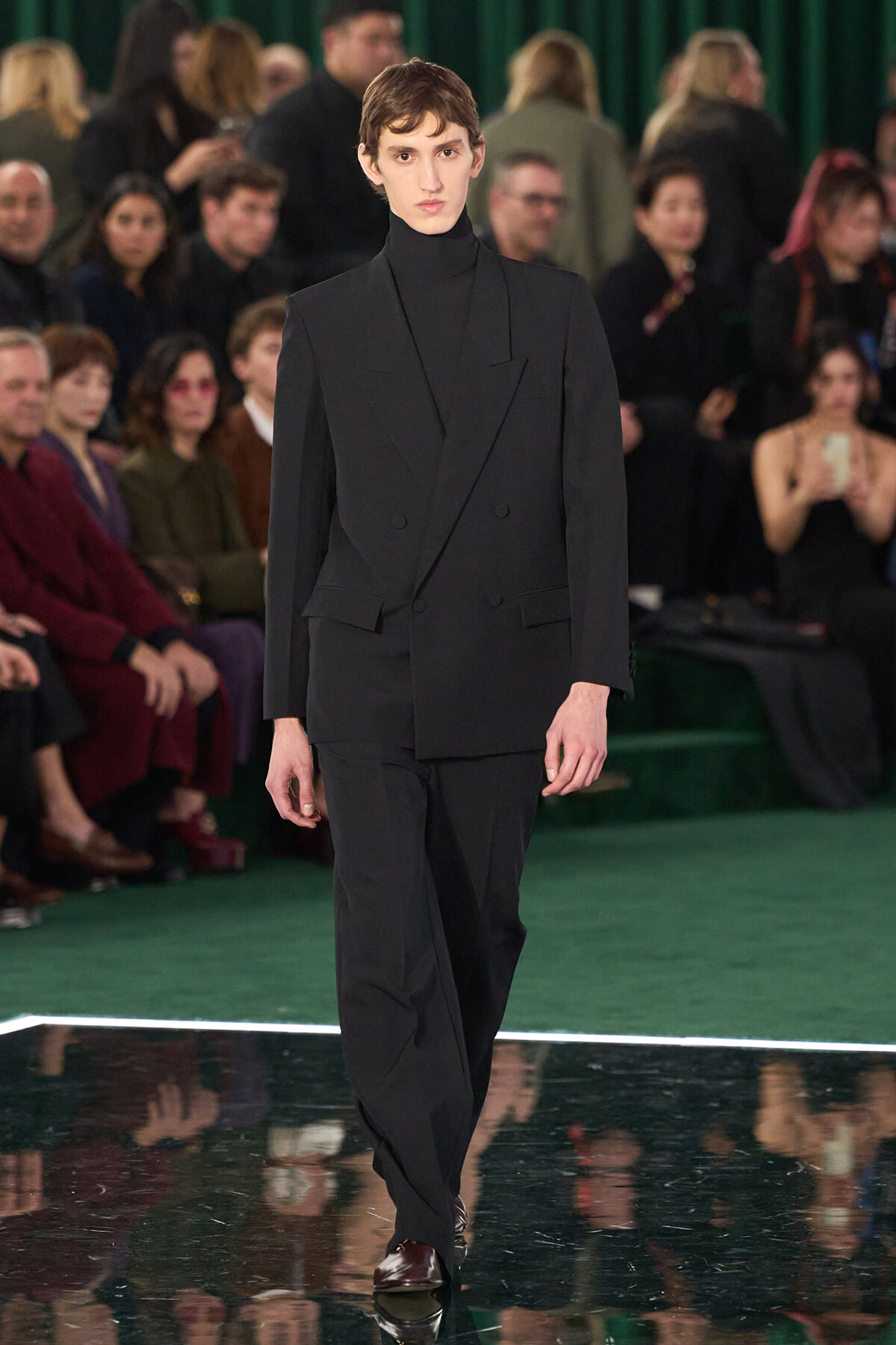 Male model in a black double-breasted suit and turtleneck walks the runway; green carpet and audience behind