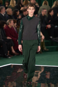 Male model walks a runway in a dark green sequined shirt, gray knit vest, and wide green trousers as seated audience watches.