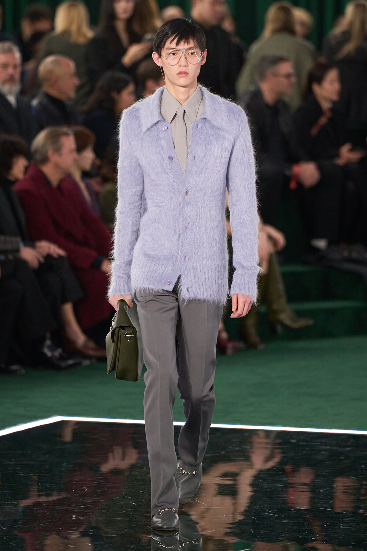 Model walking a runway in a light lavender fuzzy cardigan over a beige shirt and gray trousers, carrying a green bag at her side.