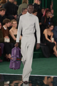 Male model on a runway wearing a pale gray turtleneck and matching wide-leg pants, carrying a purple duffel bag with green-red stripes; audience blurred in background.