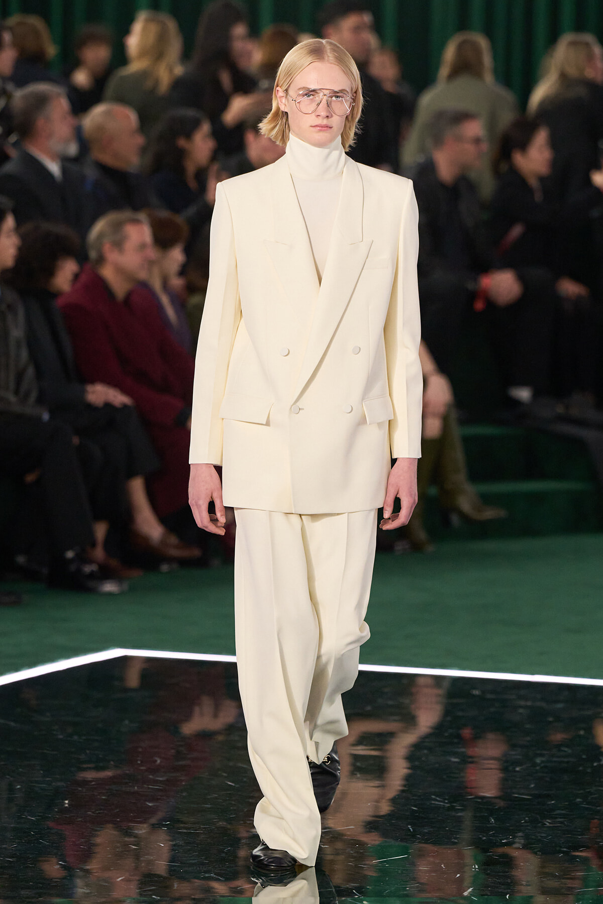 Model walking the runway in an oversized cream double-breasted suit with turtleneck and clear glasses; audience in background