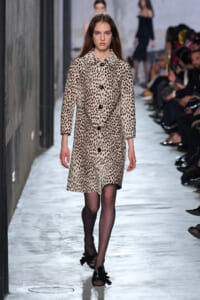 Model walks the runway wearing a knee-length beige leopard-print coat with large black buttons on a fashion show catwalk.
