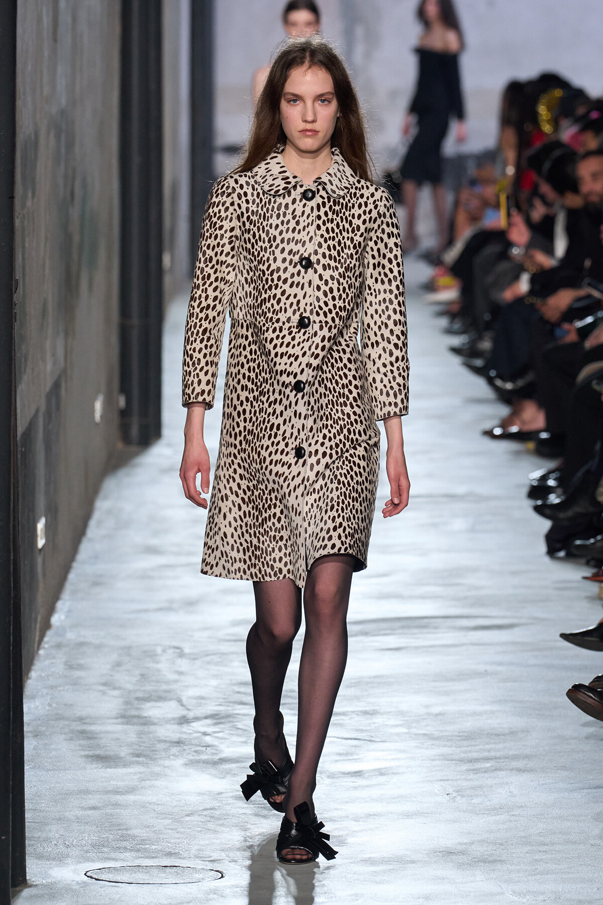 Model walks the runway wearing a knee-length beige leopard-print coat with large black buttons on a fashion show catwalk.