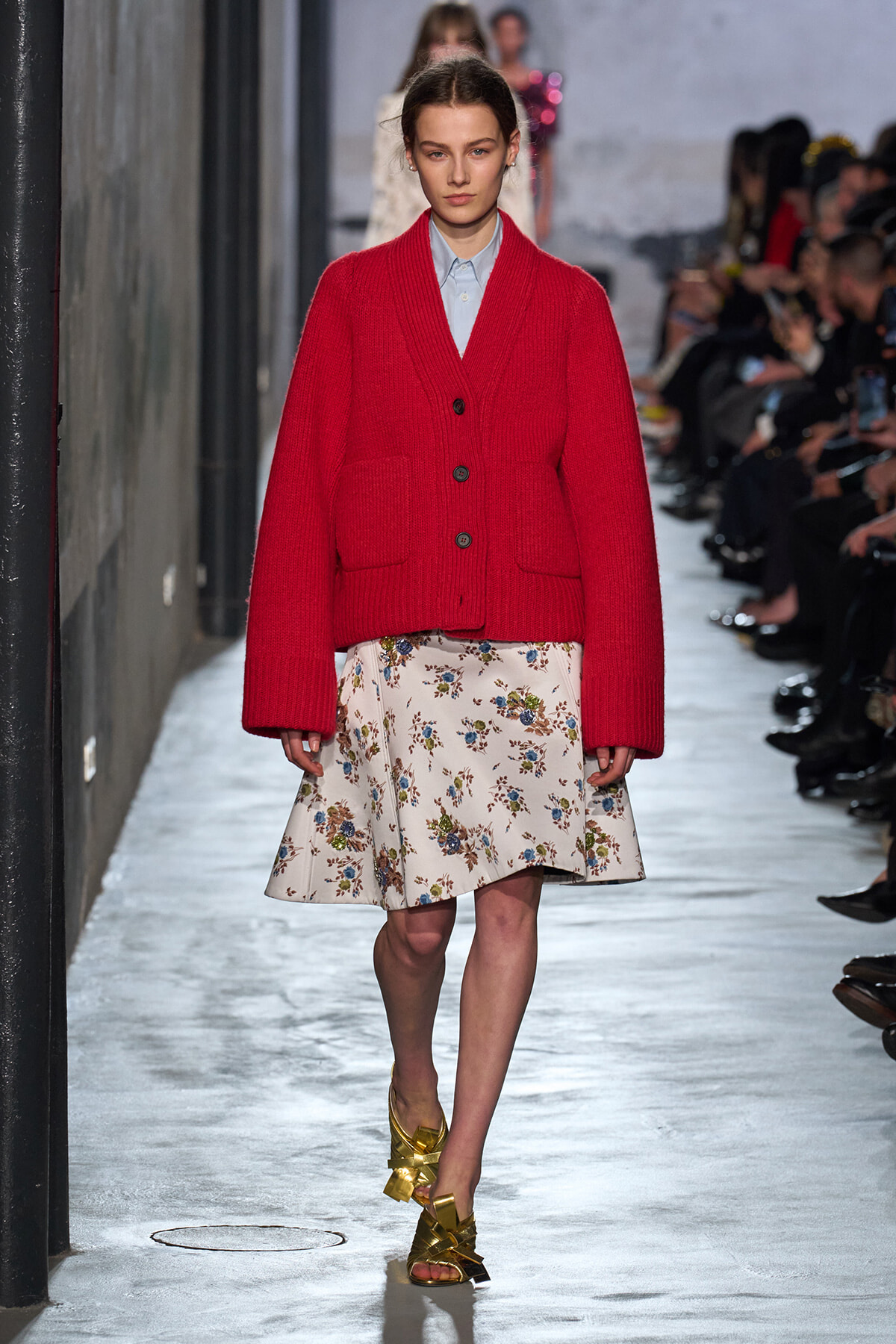Model on a runway wearing an oversized red cardigan over a light blue shirt and a white floral skirt, paired with gold sandals.