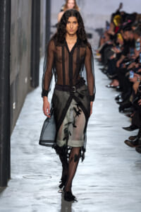 Model walking a fashion runway in a sheer black blouse and a black-and-olive sheer skirt with leaf embroidery and a belt, audience on both sides.
