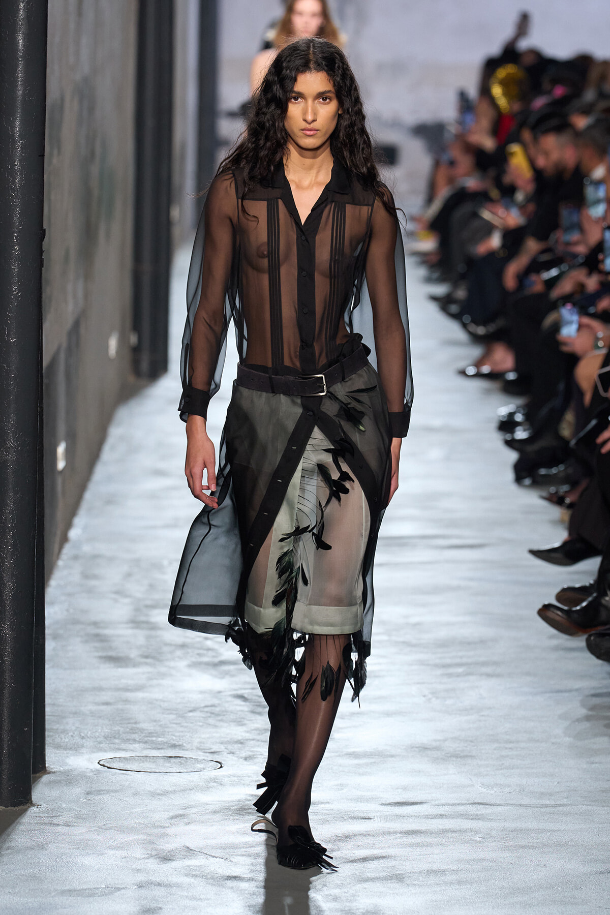 Model walking a fashion runway in a sheer black blouse and a black-and-olive sheer skirt with leaf embroidery and a belt, audience on both sides.