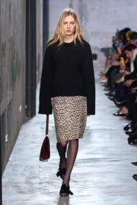 Model walking runway in a black oversized sweater and leopard-print pencil skirt, carrying a burgundy handbag.