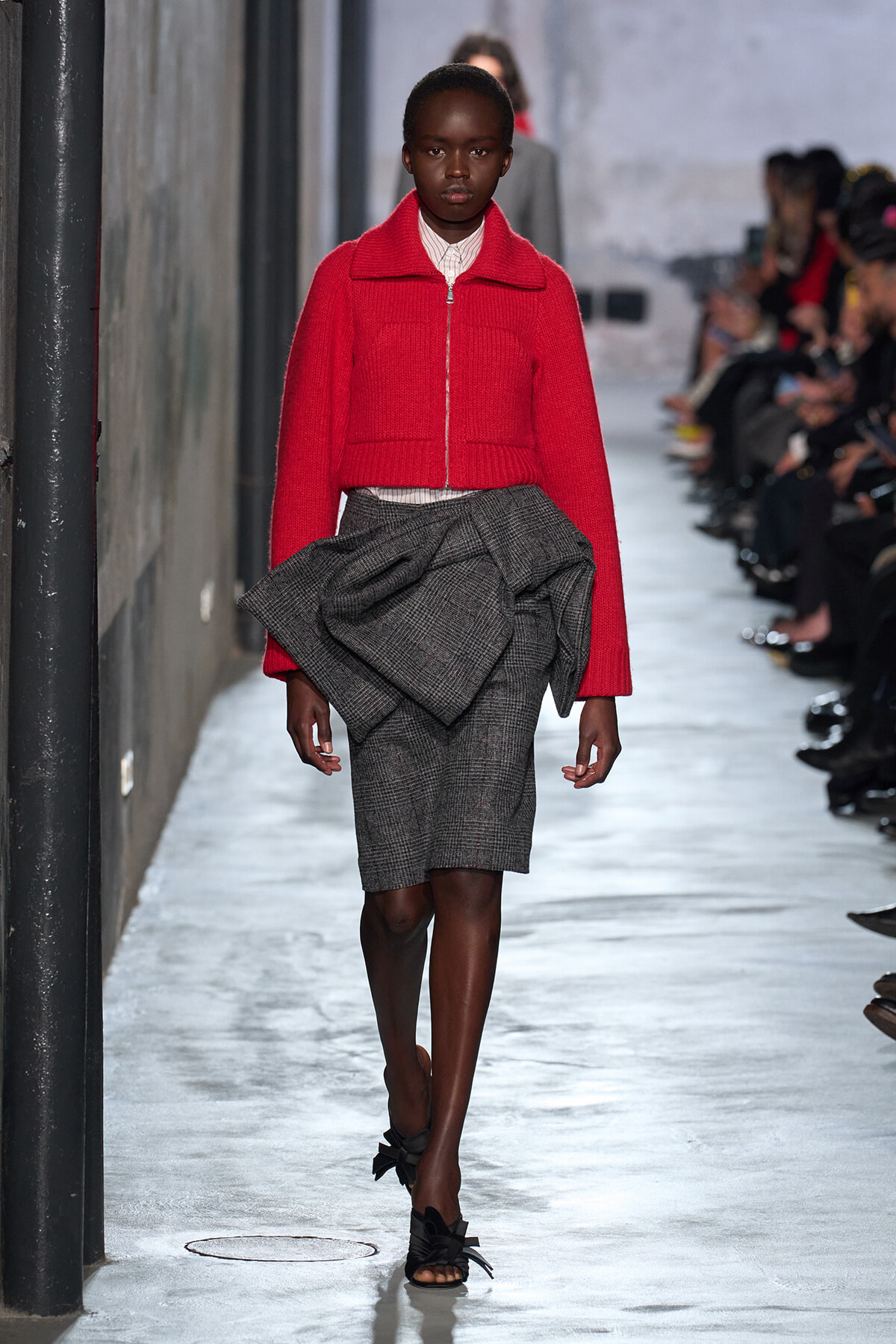 Model walks down a fashion runway wearing a bright red cropped jacket over a white top and a gray plaid knee-length skirt with a large fabric knot detail at the waist, black bow-embellished sandals.