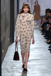 Female model walks a fashion runway in a cream floral midi dress, carrying a brown handbag and wearing black bow-strap sandals.