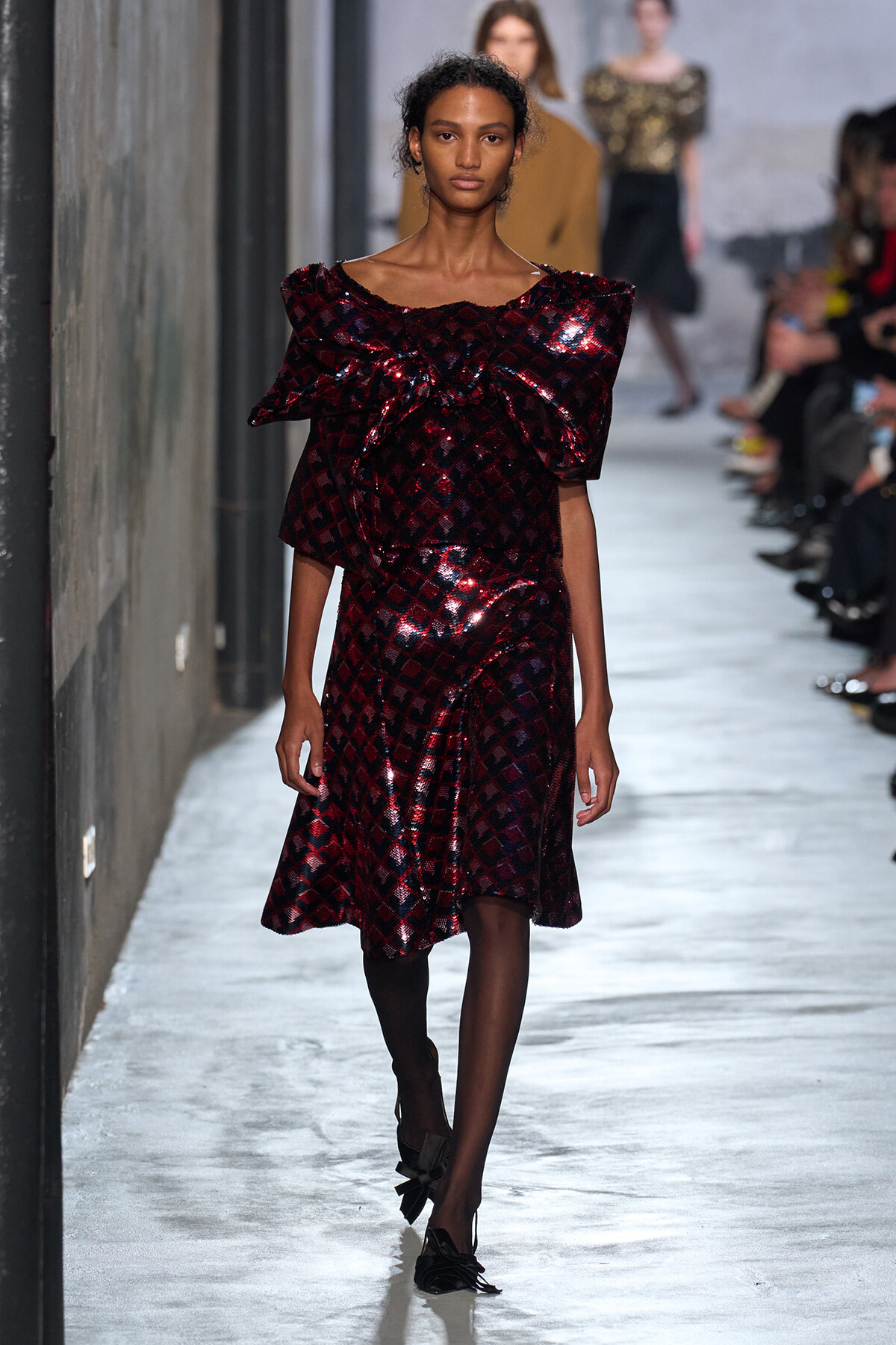 Model walking a runway in a burgundy sequin dress with a large ruffled neckline and a checkered pattern.