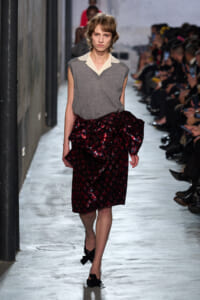 Runway model wearing a gray sleeveless top with a cream collar and a shiny red sequin plaid skirt beside the audience.