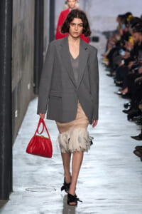 Runway model wearing a gray oversized blazer over a V-neck top and beige feathered-hem skirt, carrying a bright red handbag and black bow-embellished sandals. The scene is a fashion show runway with seated spectators.