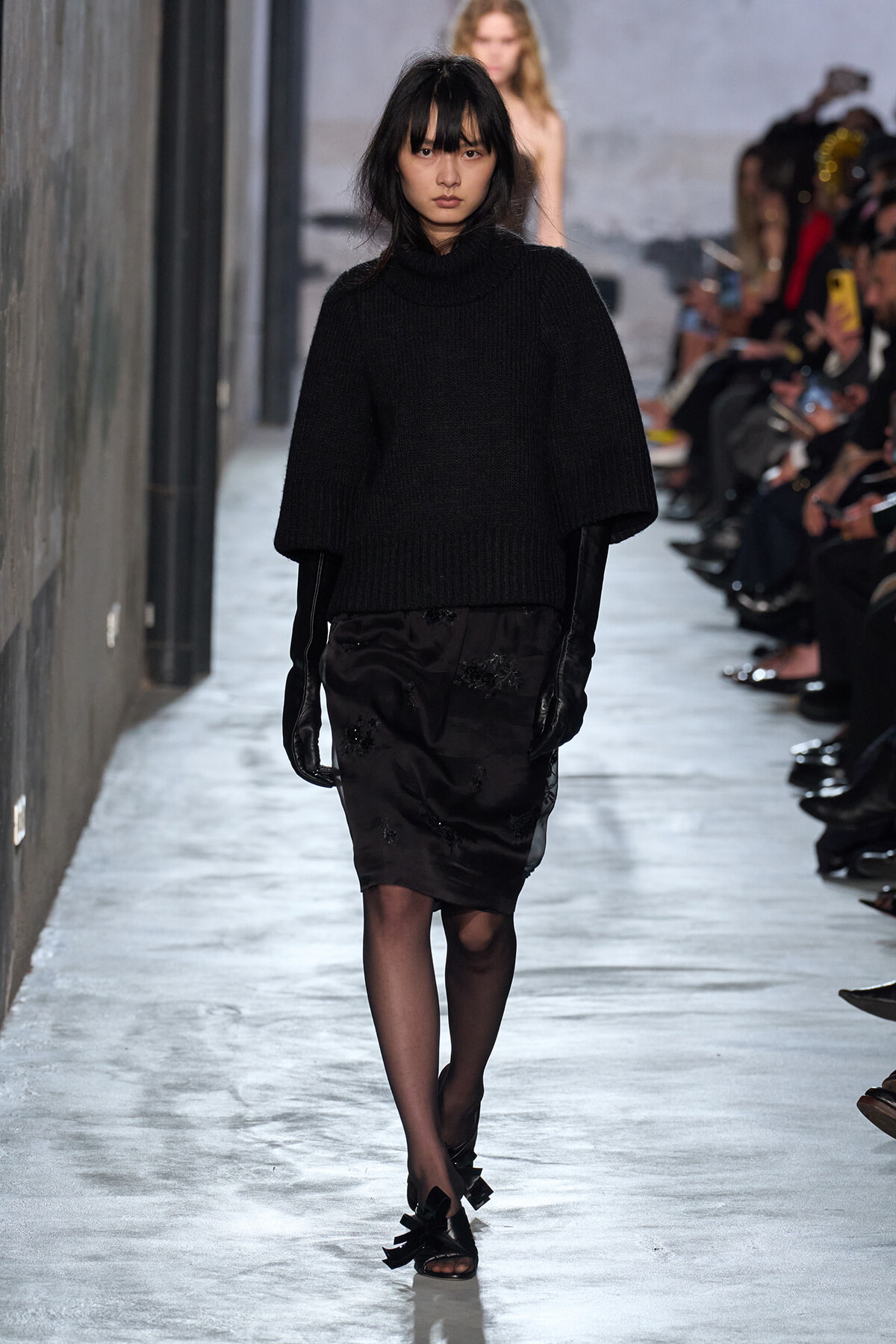 Model walking a fashion runway in an oversized black knit sweater, a black embellished knee-length skirt, sheer tights, and black bow-strap sandals, with audience along the sides.