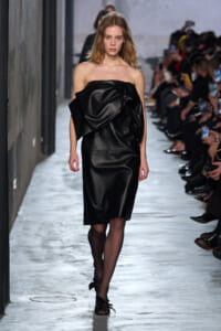 Model walks a runway in a black strapless dress with a large folded drape at the bodice, audience along the sides.