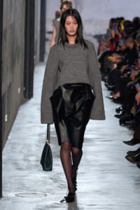 Model struts down the runway in a gray knit sweater and black leather skirt, carrying a dark green handbag as the audience watches from the sides.