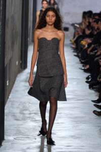 Model walks a runway in a strapless gray tweed dress with a structured bodice and layered skirt, black tights, and bow-tie flats, audience along the sides.