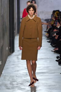 Model walking a runway in a camel coat with a matching knee-length skirt, neutral-toned outfit and sandals.