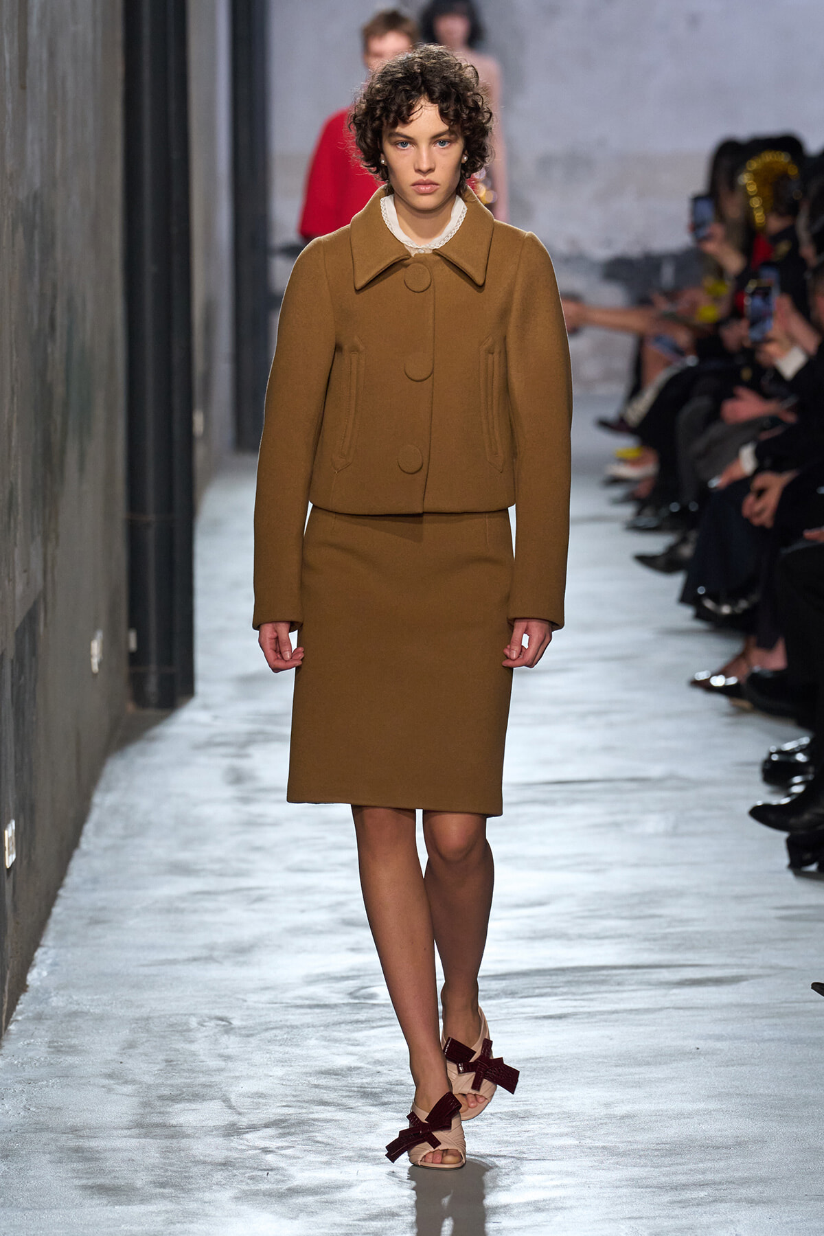Model walking a runway in a camel coat with a matching knee-length skirt, neutral-toned outfit and sandals.