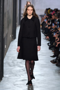 Female model walks a fashion show wearing a black knee-length coat with a large collar and round buttons, audience in background