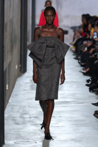 Model on the runway wearing a gray plaid off‑the‑shoulder dress with a large bow at the bodice.