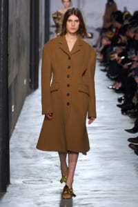 Model walking a fashion runway in a long camel-brown wool coat with black buttons and wide lapels; gold sandals visible at the feet.