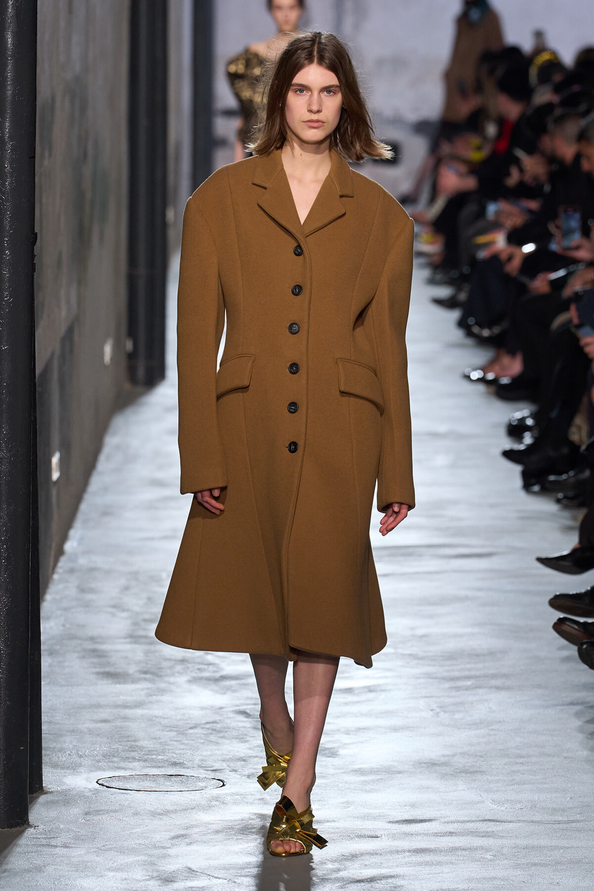 Model walking a fashion runway in a long camel-brown wool coat with black buttons and wide lapels; gold sandals visible at the feet.