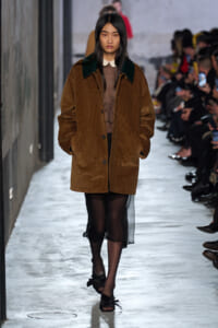 Model walks the runway in a brown oversized corduroy coat with a white collar, sheer black dress underneath, and black bow-strap sandals.
