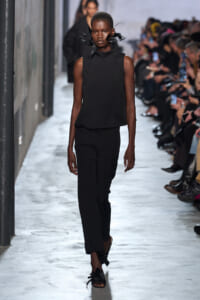 Model walks a runway in a black sleeveless top and wide-leg pants, with a dramatic black feather neckpiece.
