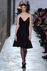 Model on a runway wearing a black strapless knee-length dress with a gathered knot at the bust.