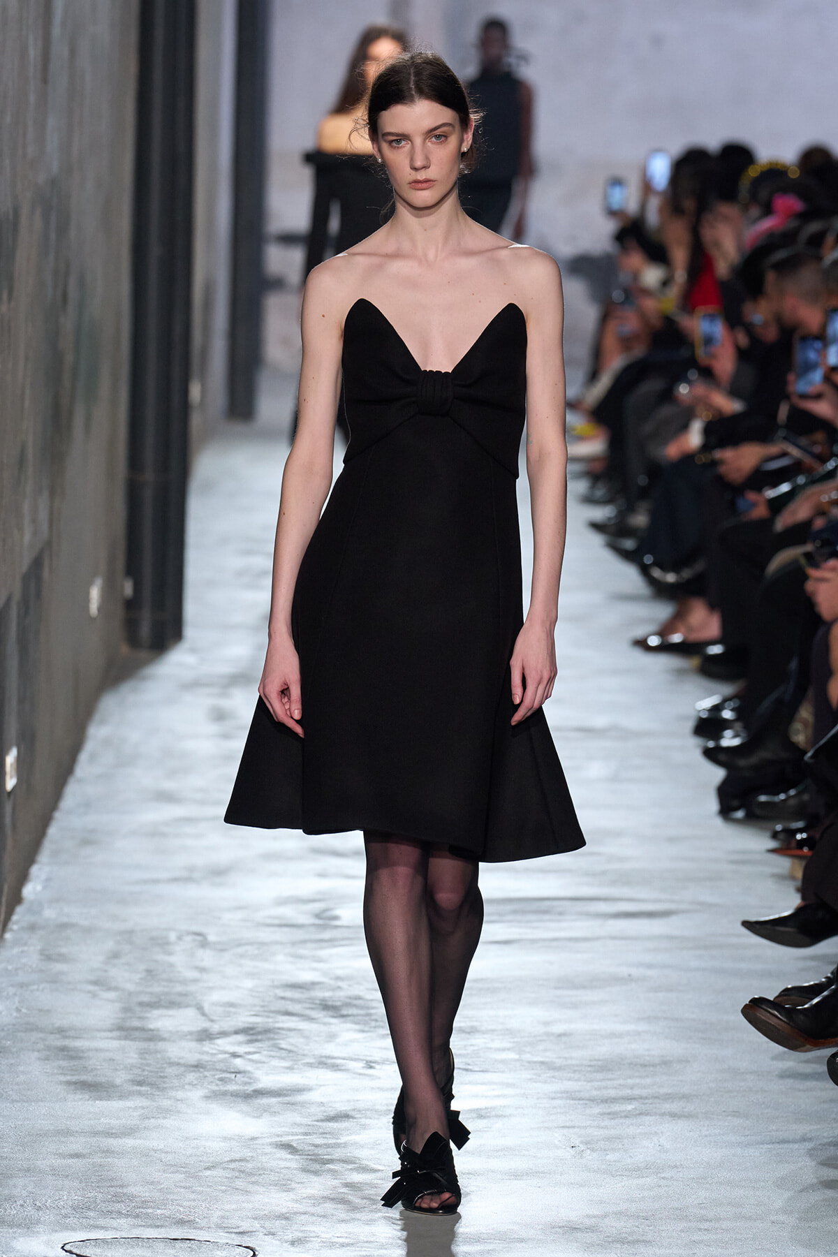 Model on a runway wearing a black strapless knee-length dress with a gathered knot at the bust.