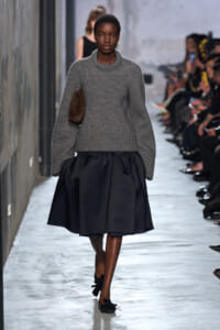 Model walking a runway in a gray chunky knit sweater and black voluminous skirt, carrying a brown clutch on her side with audience in the background.