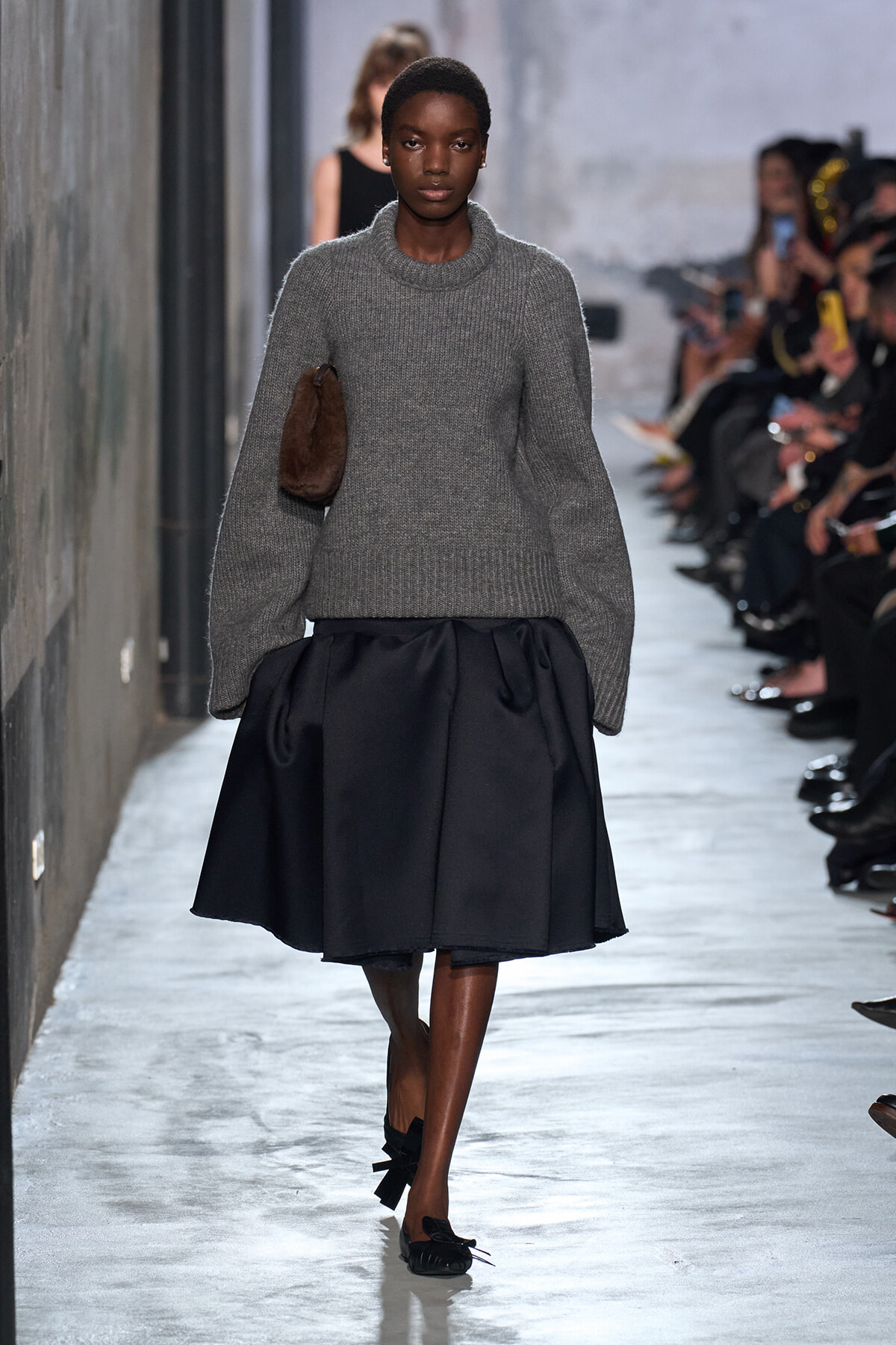 Model walking a runway in a gray chunky knit sweater and black voluminous skirt, carrying a brown clutch on her side with audience in the background.