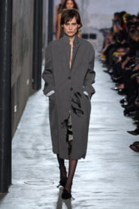 Model walks a runway wearing a long gray oversized coat with hands in pockets, audience visible along the catwalk in the background.