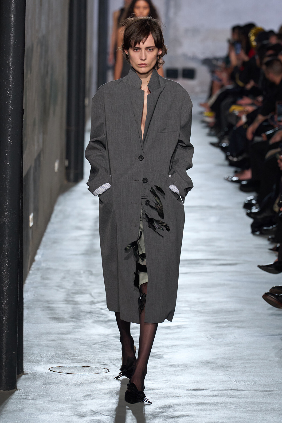 Model walks a runway wearing a long gray oversized coat with hands in pockets, audience visible along the catwalk in the background.