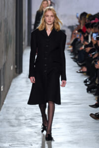 Female model walking a runway in a long black button-front coat, audience on both sides observing fashion show.