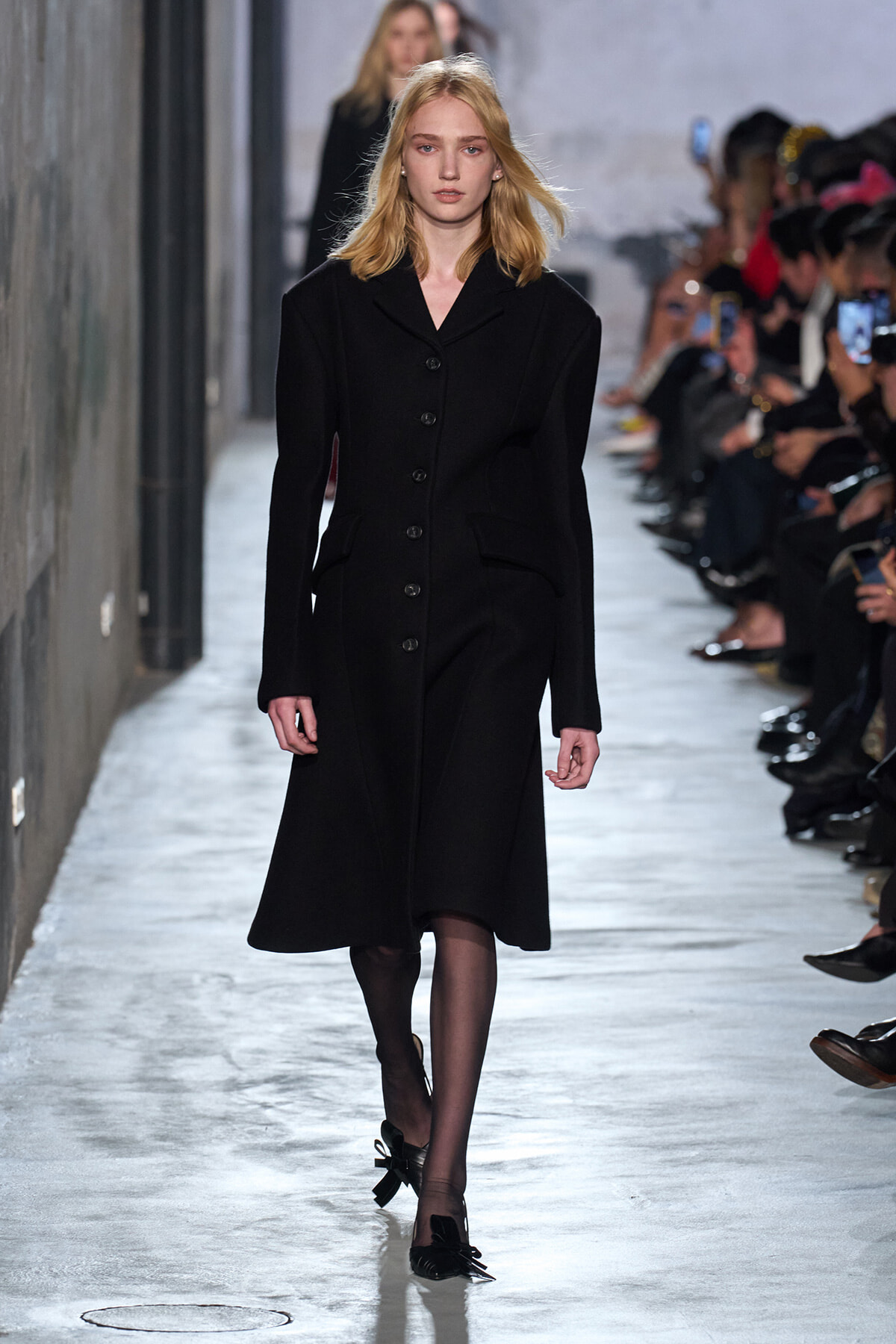 Female model walking a runway in a long black button-front coat, audience on both sides observing fashion show.