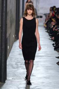 Female model walking a runway in a sleek black sleeveless sheath dress, audience on both sides.