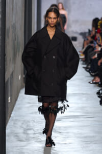 Model walks the runway in an oversized black double‑breasted coat with dramatic puff sleeves and a feathered hem at the bottom.