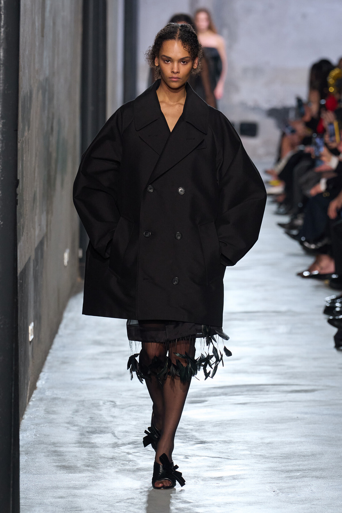 Model walks the runway in an oversized black double‑breasted coat with dramatic puff sleeves and a feathered hem at the bottom.