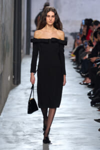 Model walks the runway in a black off-the-shoulder midi dress with a bow at the neckline, holding a small black handbag. Get audience in background is watching.