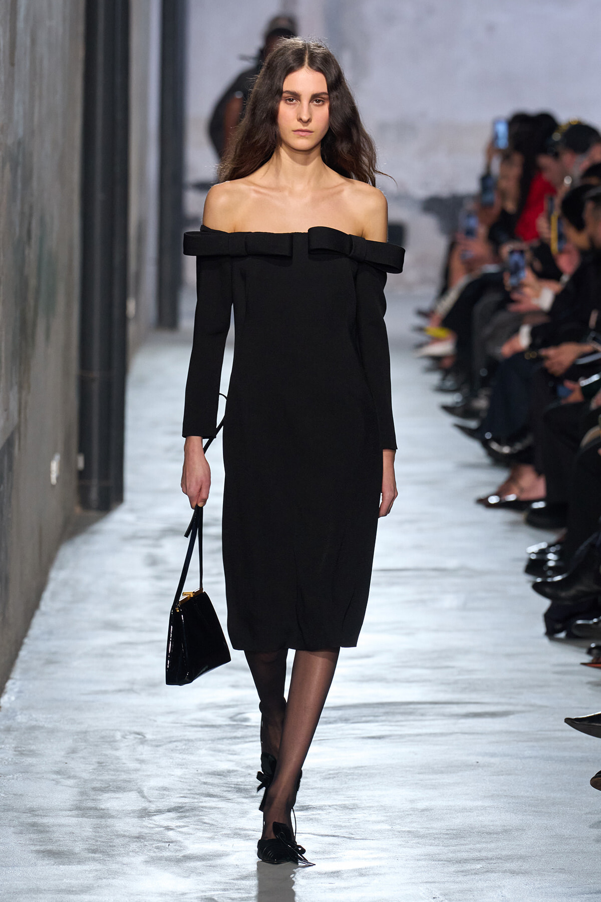 Model walks the runway in a black off-the-shoulder midi dress with a bow at the neckline, holding a small black handbag. Get audience in background is watching.