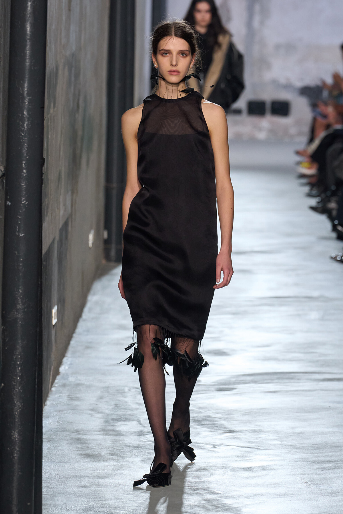 Model walks a runway in a black satin sleeveless sheath dress with a sheer yoke and feathered fringe at the hem, paired with bow-embellished heels.