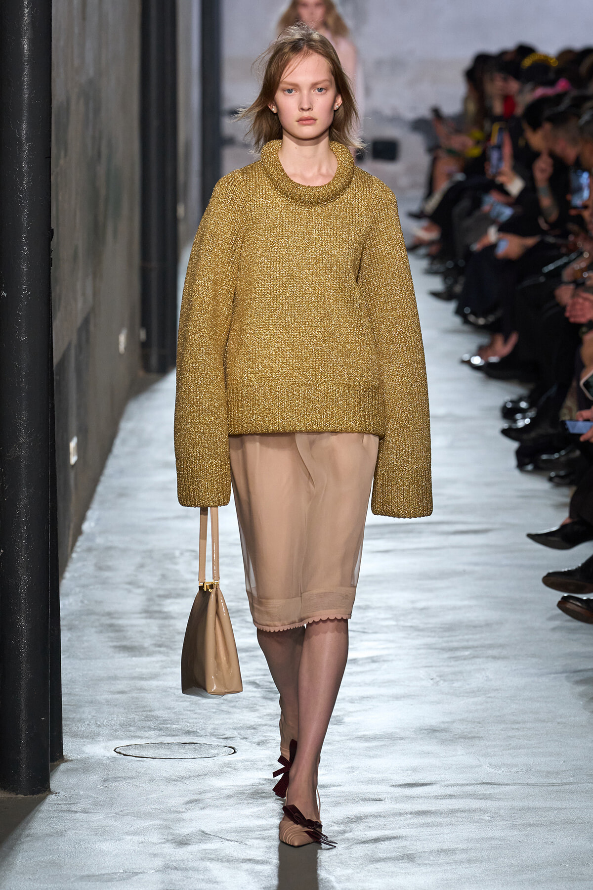 Model walking a runway in a gold chunky-knit sweater over a beige knee-length skirt, carrying a beige handbag.