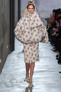 Model walks the runway in a large white floral-print coat with matching knee-length dress beneath, pastel-green bow-heeled sandals