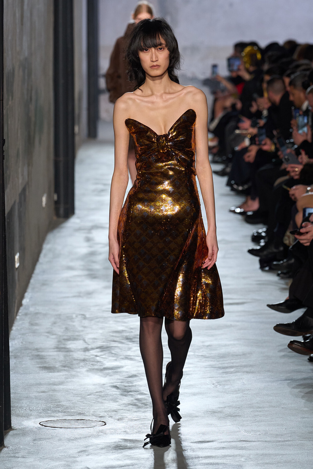 Model walks the runway in a strapless bronze sequined dress with a bow at the bust.