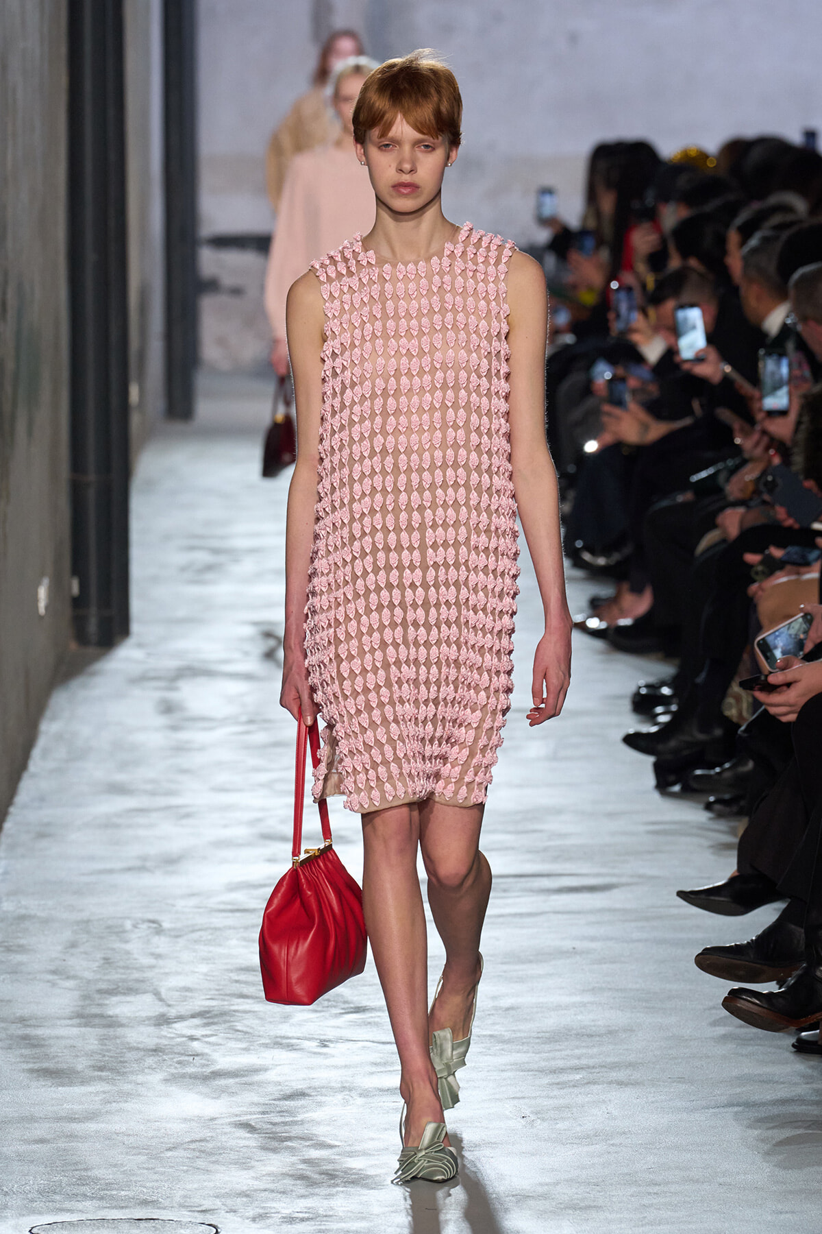 Model walks the runway in a pink textured sleeveless dress, carrying a red bag.